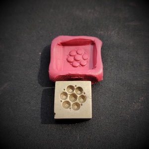 Puede incluir: Primer plano de dos moldes cuadrados, uno rojo y otro de latón. El molde rojo tiene un diseño en relieve de una flor con un círculo en el centro. El molde de latón tiene un diseño en relieve de una flor con nueve círculos.