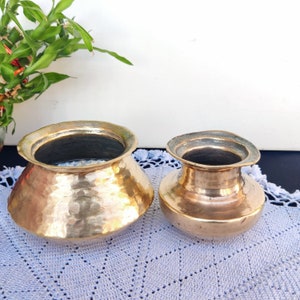 Puede incluir: Dos ollas de latón martilladas, una más grande que la otra, sobre una servilleta de ganchillo azul claro. La olla más grande tiene una abertura más ancha y un fondo redondeado. La olla más pequeña tiene una abertura más estrecha y un fondo más redondeado.