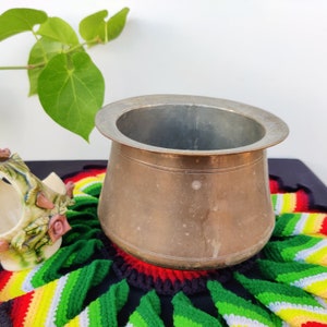 May include: A vintage brass pot with a wide, round opening and a slightly flared lip. The pot is sitting on a colorful crocheted mat.
