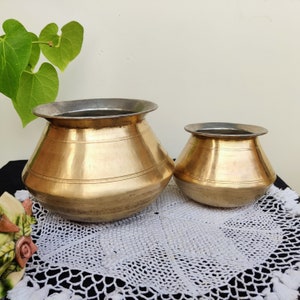 May include: Two antique brass pots, one larger than the other, are displayed on a white crocheted doily. The pots have a rounded shape and a shiny finish.