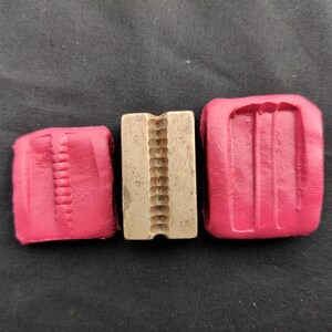 Puede incluir: Juego de herramientas de tres piezas. Una herramienta rectangular de latón con un centro ranurado está flanqueada por dos moldes rojos rectangulares cubiertos de cuero. Las herramientas están dispuestas sobre un fondo oscuro.