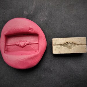 Puede incluir: Primer plano de un molde de silicona rosa con un sello de metal. El molde tiene una impresión rectangular de un diseño en forma de corazón con una banda decorativa. El sello de metal es una pieza rectangular de metal con un diseño en forma de corazón en relieve y una banda decorativa.