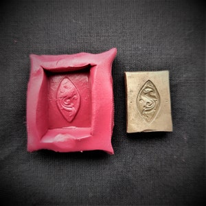 Puede incluir: Un molde de silicona rojo con un diseño en relieve de una hoja estilizada con un pájaro en su interior. Junto al molde hay un sello de latón con el mismo diseño.