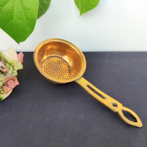 May include: A gold-colored metal tea strainer with a round bowl and a long handle. The handle has a decorative design.