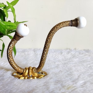 Pode incluir: Um gancho de metal dourado com um design decorativo e duas maçanetas de porcelana branca. O gancho está montado em uma superfície branca.