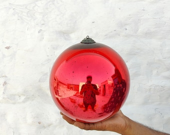 Puede incluir: Un adorno de Navidad grande, de vidrio rojo, con una superficie reflectante. El adorno es redondo y tiene una parte superior plateada. Una persona se refleja en el adorno.