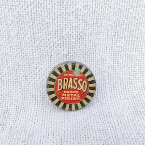 May include: A round, metal tin with a black and yellow striped design. The tin has a red label with the text "BRASSO PASTE METAL POLISH" in white lettering. The text "BRITISH MAKE" is printed in smaller white lettering above the word "BRASSO".