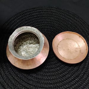 May include: A copper-colored pot with a silver-colored rim and a matching lid. The pot sits on a copper-colored base, and the lid is placed to the side. The pot and lid are on a black textured surface.