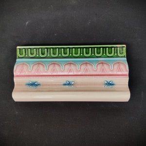 May include: A decorative ceramic tile with a pink, blue, and green floral design. The tile has a scalloped edge and a white background.