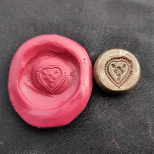 Puede incluir: Un molde de silicona rojo con un diseño en forma de corazón. El molde está al lado de un sello de metal en forma de corazón con un diseño floral.