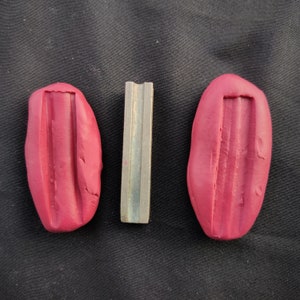 Puede incluir: Dos impresiones de arcilla roja de una barra de metal con una sección transversal rectangular. La barra de metal está en el centro de la imagen.