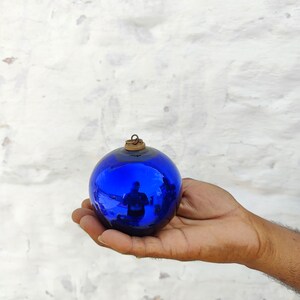 May include: A deep blue glass bauble, a Christmas tree decoration. The spherical ornament has a reflective surface and a small, aged metal cap. The bauble is held in a hand against a white textured background. A classic festive decoration.
