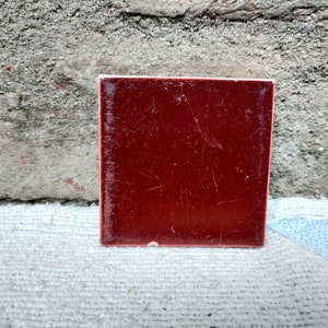 May include: A square tile with a deep red glaze. The tile has a slightly textured surface and is slightly worn.