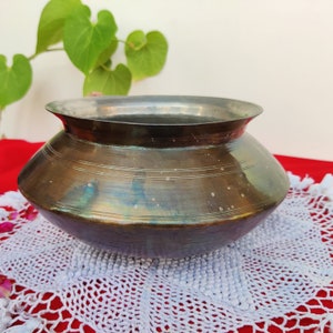 May include: A vintage, dark brown metal bowl with a slightly flared rim. The bowl has a worn, aged patina and is sitting on a white crocheted doily.