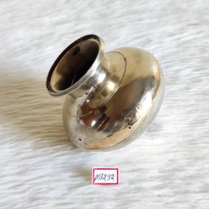 May include: A small, round, brass pot with a wide opening. The pot has a smooth, shiny surface and a slightly dented bottom. The pot is sitting on a white surface.