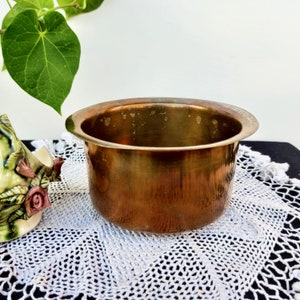 Puede incluir: Un macetero redondo de latón con acabado martillado. El macetero está sobre un encaje blanco de ganchillo.