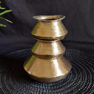 Puede incluir: Una pila de tres ollas antiguas de latón. Las ollas tienen una textura martillada y un acabado dorado brillante. La olla más grande está en la parte inferior, la más pequeña en la parte superior. Las ollas probablemente se usan para cocinar.