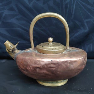 May include: A copper and brass teapot with a hammered copper body and a brass handle and lid. The teapot has a spout shaped like a bird's head.