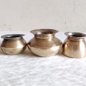 May include: Three antique brass pots with a silver-coloured rim. The pots are different sizes and have a rounded shape. The brass has a warm, golden hue, and the pots are displayed on a white surface.