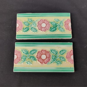 Op de afbeelding: Twee vintage keramische tegels met een bloemenmotief. De tegels zijn geel met groene bladeren en roze bloemen met witte centra. De tegels hebben een groene rand.