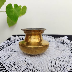 Puede incluir: Una pequeña olla de latón martillado con una base ancha y redonda y un borde ligeramente acampanado. La olla está sentada sobre un encaje blanco de ganchillo.