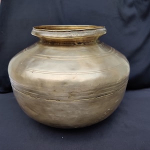 May include: A vintage brass pot with a rounded body and a slightly flared rim. The pot features horizontal etched lines around its circumference, adding a decorative element to its aged, metallic surface. The pot has a tarnished appearance.