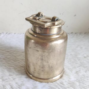 Puede incluir: Un recipiente de latón antiguo con tapa abisagrada. El cuerpo cilíndrico tiene una pátina ligeramente envejecida y la tapa presenta un asa rectangular. El recipiente se utiliza probablemente para almacenamiento o exhibición. El diseño general es sencillo y funcional.