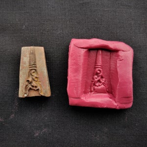 Puede incluir: Un primer plano de un molde de metal para hacer joyas. El molde está hecho de metal oscuro y tiene un diseño detallado. El molde está sobre una superficie oscura. El molde está al lado de un molde de silicona rojo.