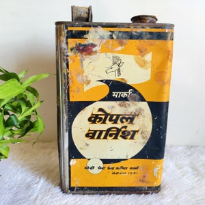 May include: A vintage metal tin of Kopal Varnish, a type of wood finish. The tin is yellow and black with a deer logo and the text "माकी कोपल वार्निश" in Hindi. The tin is rusty and shows signs of wear.