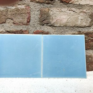Può includere: Due piastrelle quadrate in ceramica azzurra. Le piastrelle sono affiancate, con una finitura liscia e lucida. Lo sfondo presenta un muro di mattoni ruvido e strutturato.