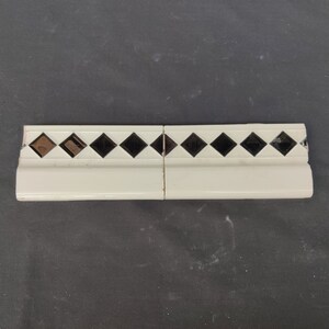 May include: Two white ceramic tile pieces with a black diamond pattern border. The tiles are designed for use as a decorative trim.