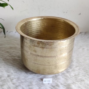 May include: A hammered brass pot with a wide, rounded rim and a cylindrical body. The pot has a textured surface and a warm, golden color. The pot is sitting on a white surface. The text "ME 441" is on a small white label.