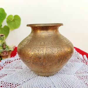 May include: A brass vase with intricate engraved designs, featuring a traditional Indian pattern. The vase is sitting on a white crocheted doily.