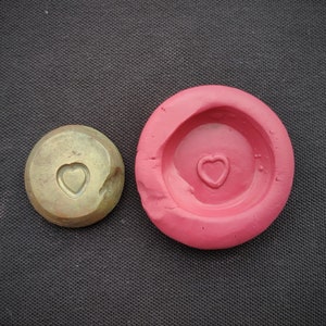 Puede incluir: Primer plano de dos moldes redondos, uno de latón con un diseño en forma de corazón y el otro de silicona roja con una hendidura en forma de corazón.