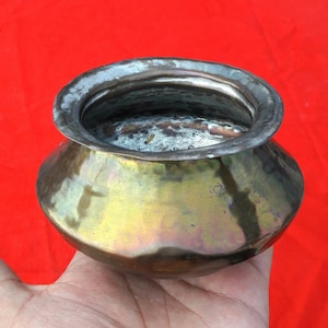 May include: A small, round, brass pot with a worn, aged patina. The pot has a wide, rounded base and a narrow opening. The inside of the pot is visible and appears to be clean.