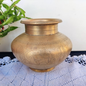 May include: A brass vase with intricate etched designs, including an elephant, on a light blue crocheted surface.