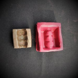 Puede incluir: Primer plano de dos moldes para hacer jabón o velas. El molde de la izquierda está hecho de metal y tiene una pátina marrón. El molde de la derecha está hecho de silicona roja y tiene una forma rectangular.
