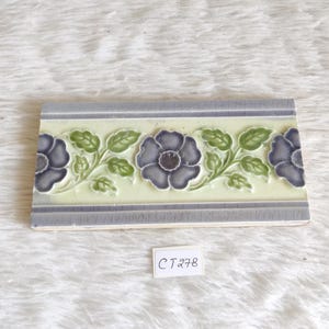 Pode incluir: Um azulejo de cerâmica vintage com um design floral. O azulejo apresenta um padrão repetitivo de flores roxas com folhas verdes sobre um fundo branco. O azulejo tem uma borda azul clara.