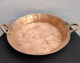 May include: A large, round copper pan with two handles. The pan has a worn, aged appearance and is likely antique.