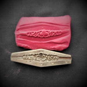 May include: A close-up of a metal mould with a detailed floral design. The mould is used to create a decorative border for jewellery or other crafts. The mould is silver in colour and has a smooth, polished finish.