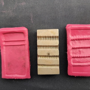 Puede incluir: Tres moldes rectangulares, dos son de silicona roja y uno es de metal plateado. El molde de metal tiene una superficie texturizada con líneas en relieve.