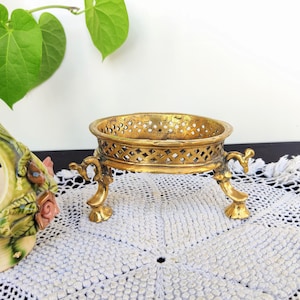 Op de afbeelding: Een decoratieve standaard van goudkleurig metaal met een ronde bovenkant en drie poten. De standaard heeft een tralieontwerp en is perfect voor het presenteren van kleine voorwerpen.