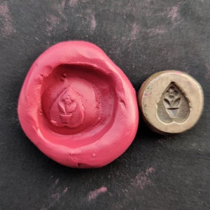May include: A close-up of a red clay impression of a heart-shaped design with a flower inside. The design is made with a metal stamp, which is also shown in the image.
