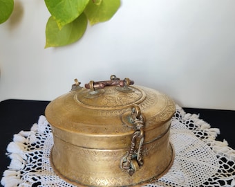 Puede incluir: Una caja redonda de latón con un diseño ornamentado y una tapa con bisagras. La caja tiene una manija decorativa y un pestillo.