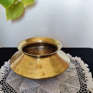May include: A round, brass pot with a wide, flared rim. The pot has a hammered finish and is sitting on a white crocheted doily.