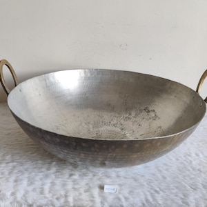 Può includere: Un grande wok in metallo martellato con manici in ottone. Il wok ha una forma a ciotola ampia e poco profonda ed è realizzato in metallo color argento. I manici sono circolari e attaccati ai lati del wok. Il wok viene probabilmente utilizzato per cucinare.