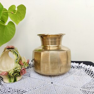 May include: A gold brass vase with a rounded body and a flared top. The vase has a geometric pattern and is sitting on a white crocheted doily.