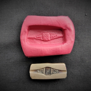 Puede incluir: Un molde de silicona rosa con una impresión detallada de un anillo con diseños intrincados. El molde está al lado de un sello de anillo de metal con el mismo diseño.