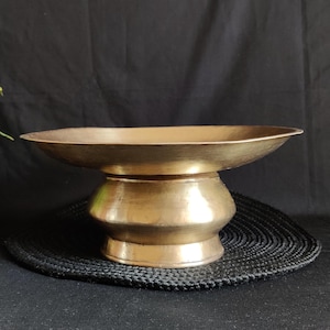 Puede incluir: Un cuenco con pedestal de color bronce con un plato ancho y poco profundo y una base redondeada. El cuenco se asienta sobre una estera negra tejida sobre un fondo oscuro. La superficie del cuenco tiene un aspecto ligeramente envejecido.