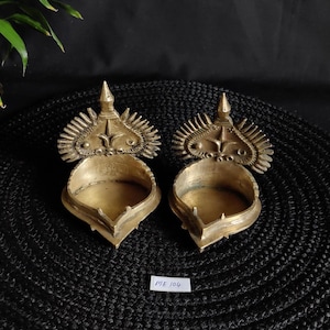 Può includere: Due antiche lampade a olio in ottone con disegni ornati a forma di raggio di sole sopra le ciotole. Le lampade hanno una parte superiore a punta e una base decorativa. Le lampade sono su una superficie nera strutturata. L'etichetta recita "ME 104".